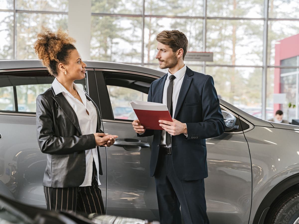 A salesman with a folder and a customer beside a car on the showroom floor.