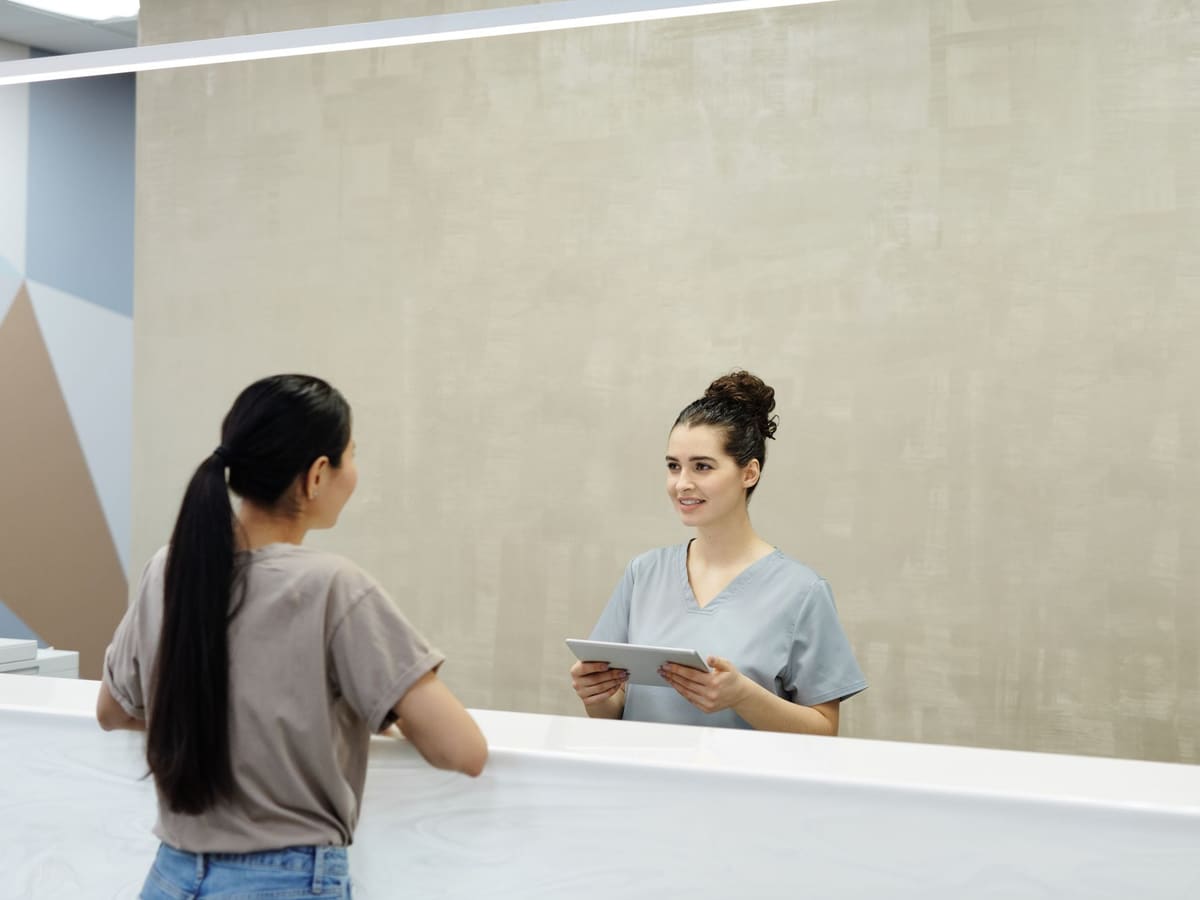 A receptionist with a tablet, smiling at a patient across the front desk.