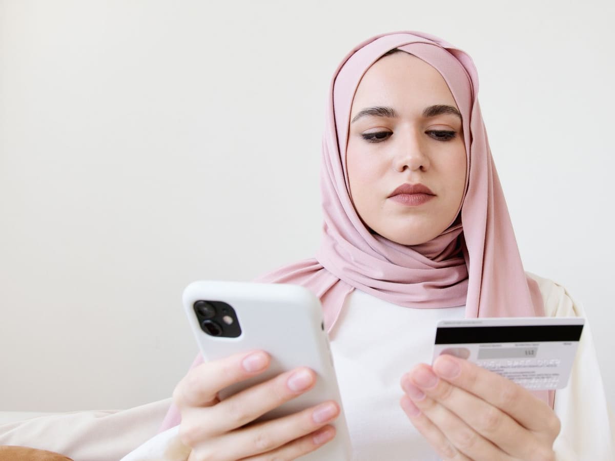 A woman in a pink headscarf, phone in one hand and a bank card in the other.