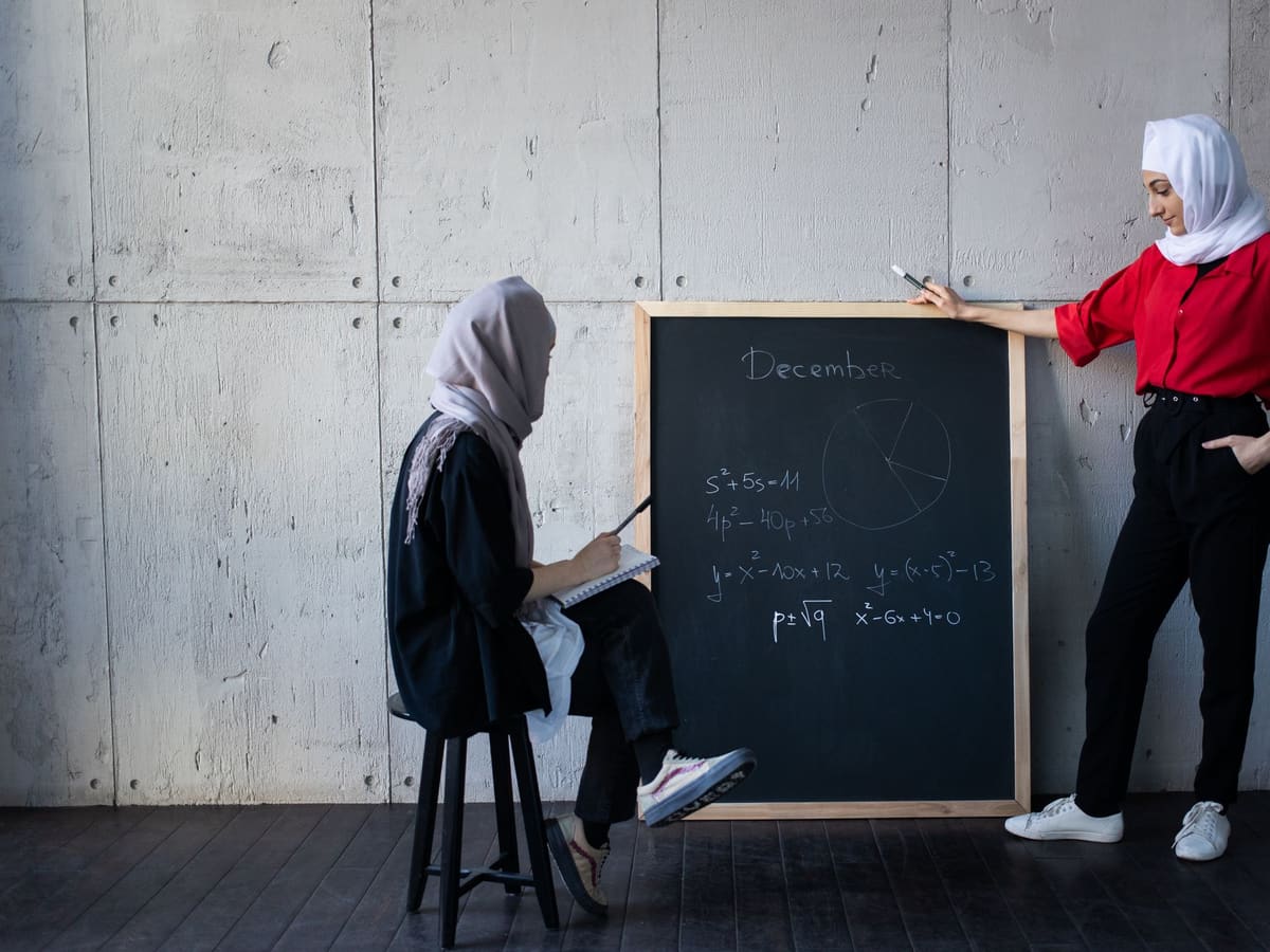 A teacher at a chalkboard covered in equations, a student on a stool taking notes.