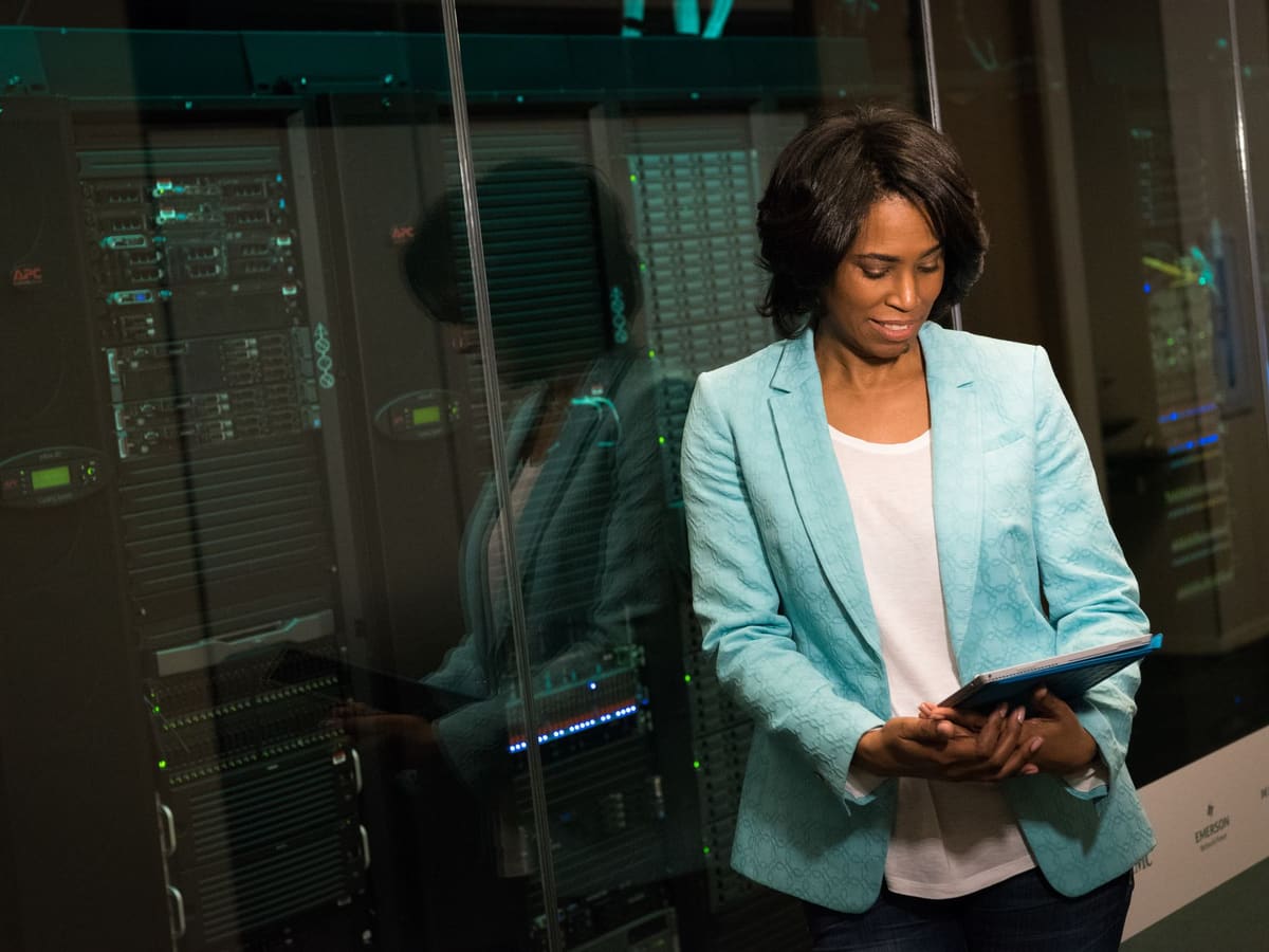 An engineer in a teal jacket reading a tablet beside rows of servers.