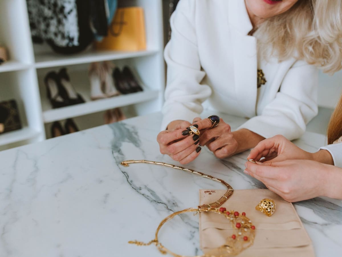 Two women at a marble table, trying on gold rings and necklaces.